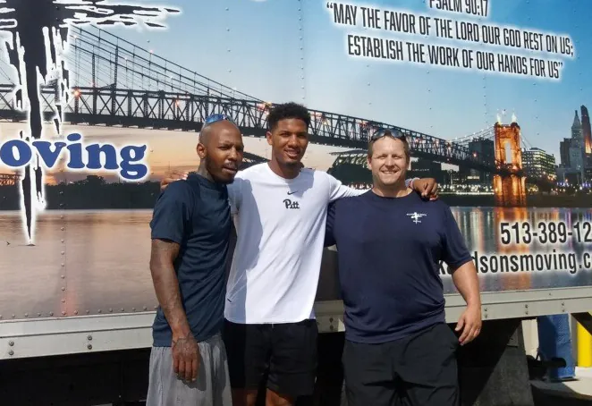 Three Clark and Sons movers standing in front of a company truck painted with a Bible verse and the Cincinnati riverfront