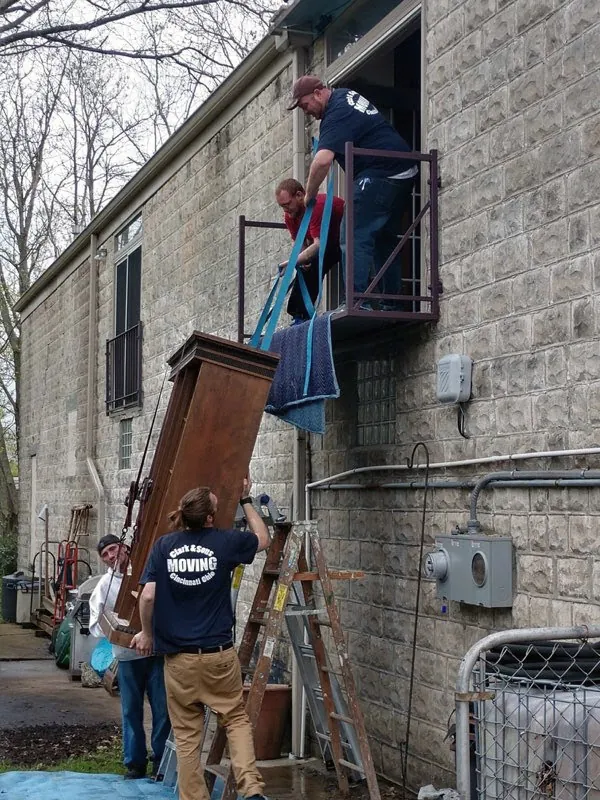 Crew lowering a tall wardrobe from a second-floor window on ropes